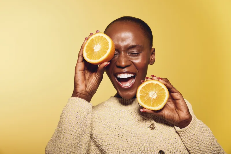 Happy African woman with dark skin holds two halves of an orange fruit near her face, smiling with closed eyes. Bright yellow background and beige sweater create warm cheerful mood.
