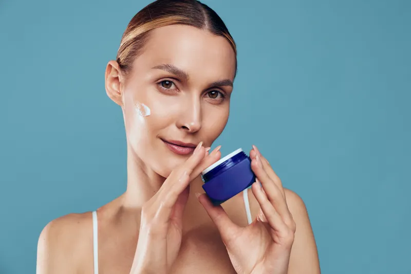 Young woman applies face cream on cheek holds blue jar skincare product looks at camera with gentle smile isolated on blue background natural beauty and skin care concept.