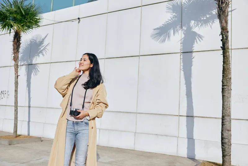 Woman with camera smiles beside a light wall and tall palm trees, wearing a long beige coat and jeans, capturing street photography in bright outdoor daylight, casual urban style and cheerful mood
