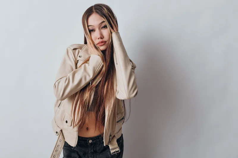 Young woman with long hair wearing a light jacket and black jeans poses on white background with hands touching her face in casual fashion style portrait.