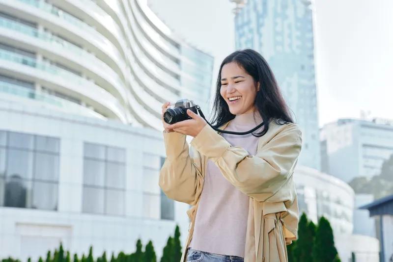 Candid shot of a smiling woman photographer outdoors in an urban setting, camera in hand, light jacket, modern buildings in background, travel and creative photography vibe.