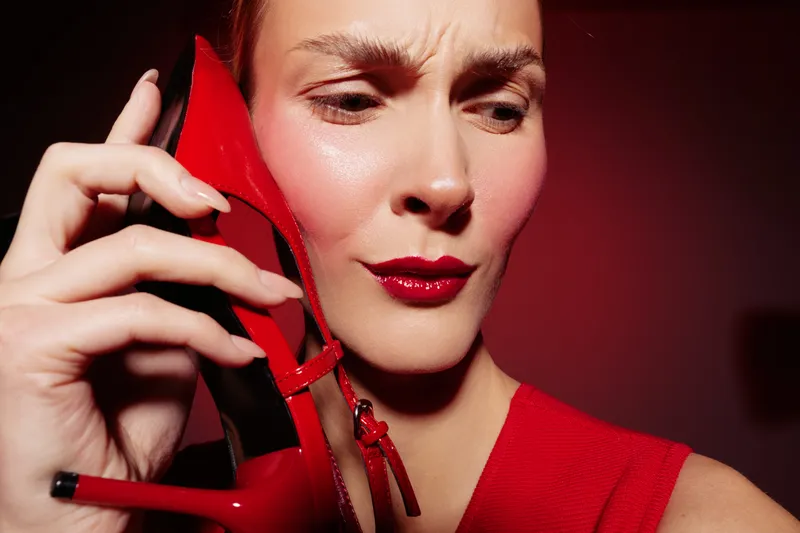 Woman in red dress holds red high heel shoe near face with confused expression. Studio portrait with dramatic lighting on red background. Concept for fashion, style, and emotions.