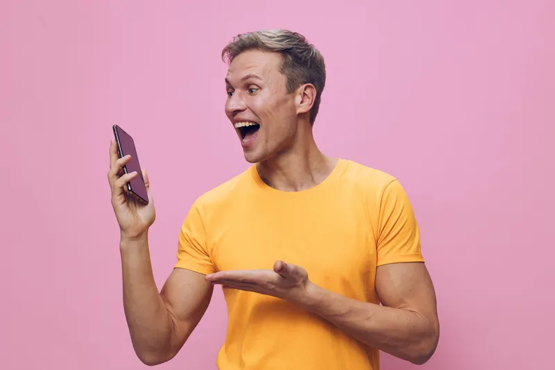 Smiling young man with short brown hair wearing bright yellow T-shirt holding smartphone in hand look excited express joy on pink background studio portrait. People lifestyle concept