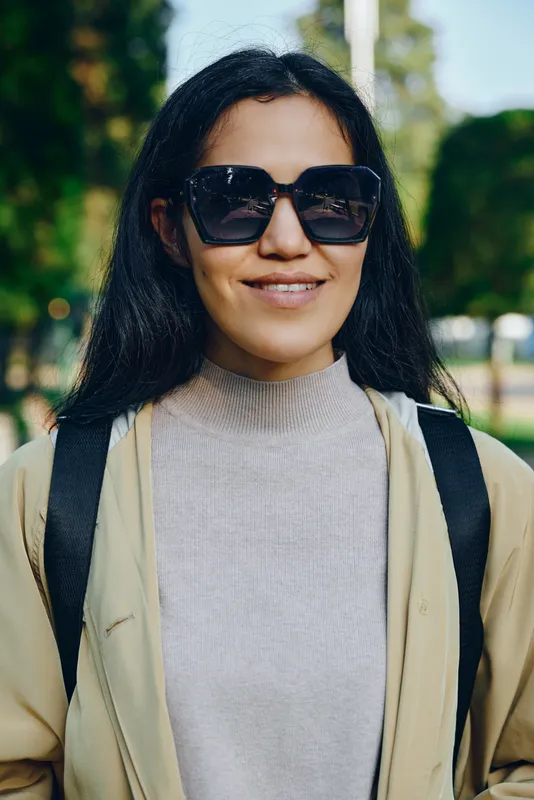 Woman wearing sunglasses and a casual beige jacket in a sunny park, smiling while carrying backpack straps, showcasing relaxed outdoor fashion and everyday style
