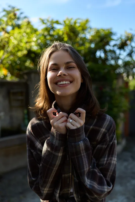 A cheerful woman in a plaid shirt outdoors, smiling at the camera in a sunny yard. Casual lifestyle portrait capturing happiness, natural light, and relaxed mood