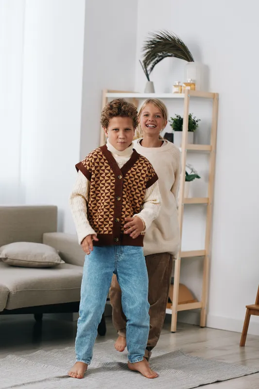 Two children stand indoors, a playful boy in a patterned knit vest and jeans at the front, smiling confidently as warm daylight fills a cozy living room.