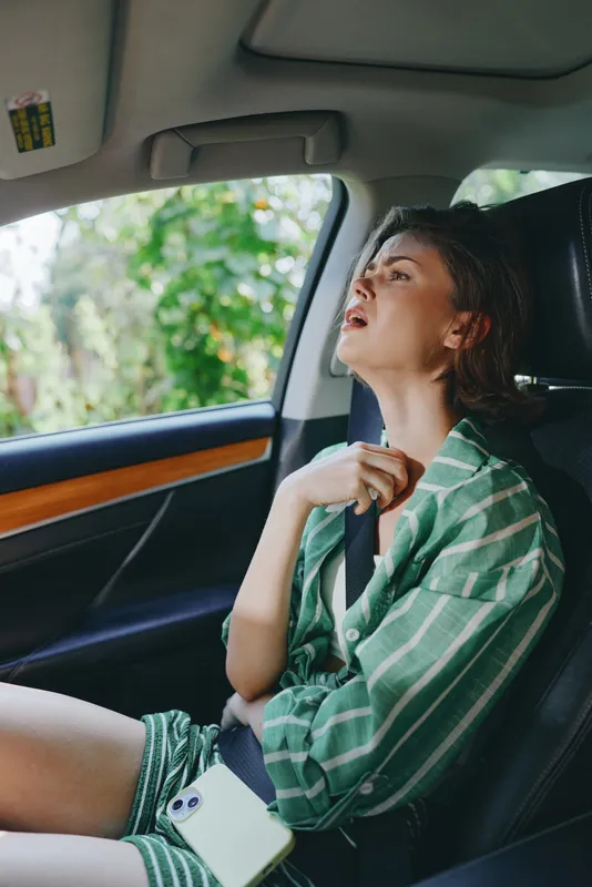 A woman sits in the driver's seat wearing a striped green shirt, hands on the wheel, looking upward with a calm, hopeful expression.