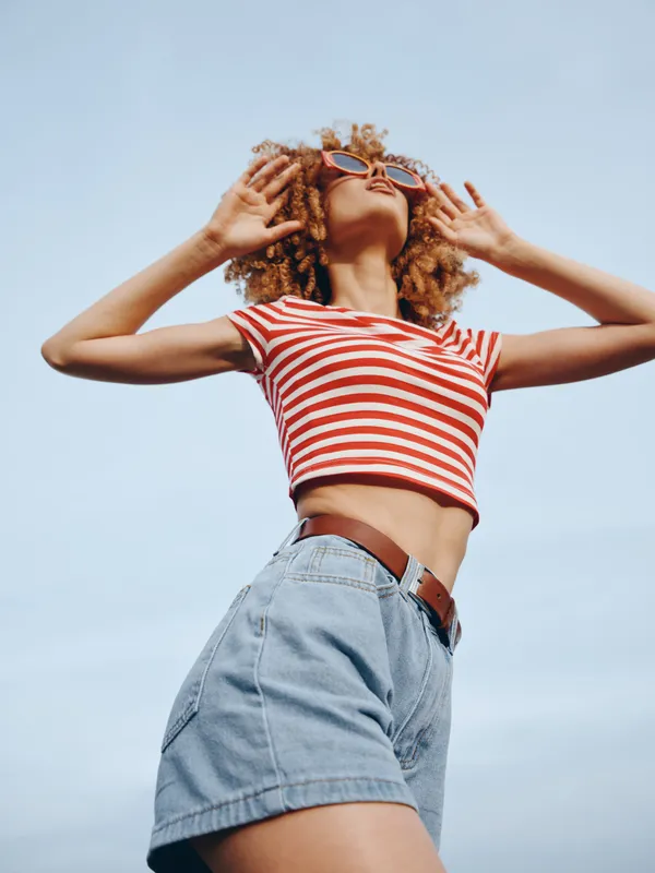 Vibrant portrait of a carefree young woman outdoors under a clear blue sky, wearing a striped crop top and denim shorts, arms raised in sunny energy.