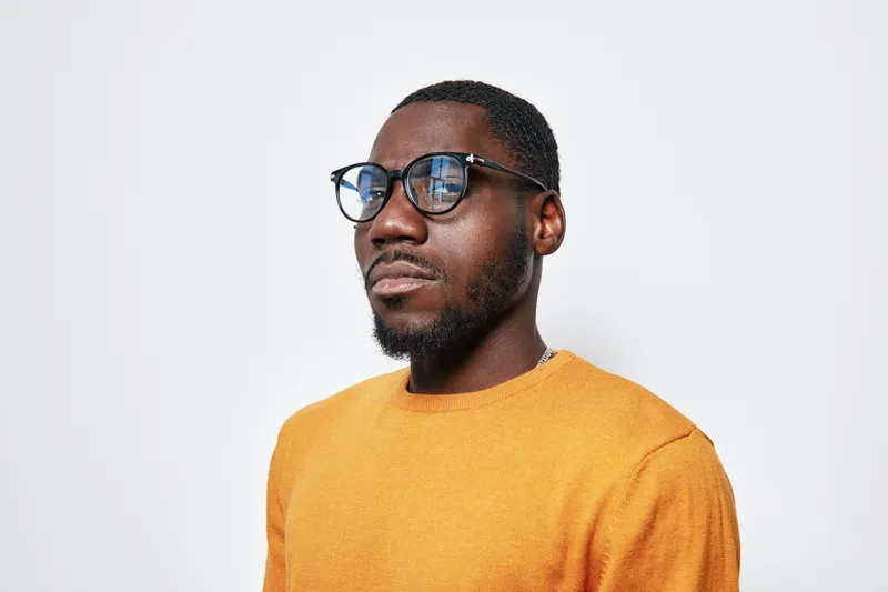 African American man with dark skin wearing glasses and orange sweater looking confident against white background. Portrait of young black male with thoughtful expression and casual style.