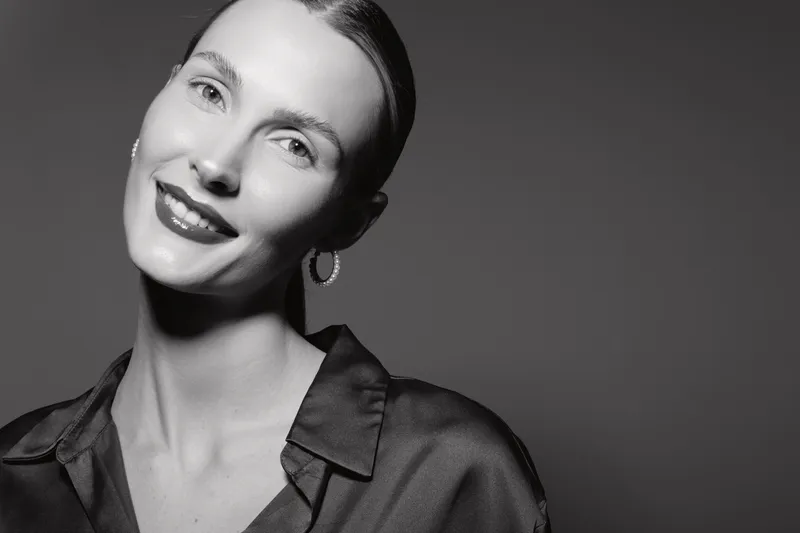 Portrait of a smiling white woman with light skin and slicked back hair wearing a dark blouse and hoop earrings on grey background.