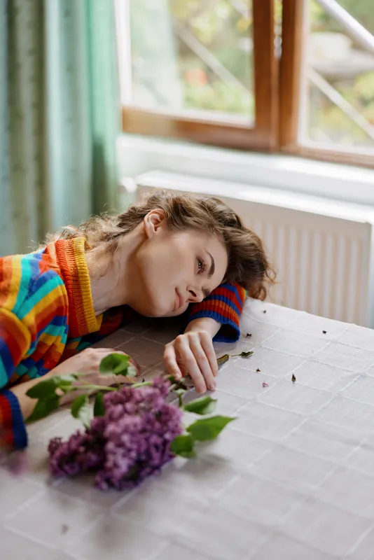 A sleeping woman with curly hair rests her head on a white table in a bright room, wearing a colorful striped sweater as a bouquet of lilac lies nearby, creating a calm, cozy scene