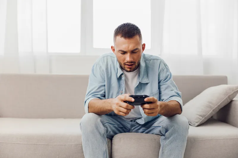 Focused man enjoying a gaming experience indoors, dressed casually in denim and a t-shirt, surrounded by a bright, minimalistic living room ambiance.
