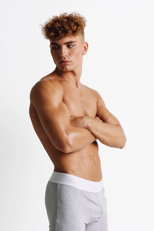 Young muscular man with curly hair standing shirtless in grey pants on white background showing defined abs and looking to side with arms crossed for body fitness presentation.