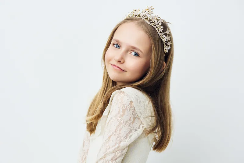 Little girl in a graceful dress with a crown, smiling softly against a light background, embodying innocence and joy in a serene portrait