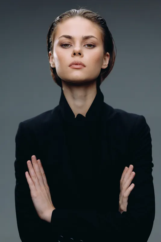 Confident woman in black jacket with folded arms posing against dark background, strong expression, professional portrait
