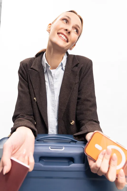Young woman in casual blazer holding passport and smartphone leans on blue suitcase, smiling and looking up. Concept of travel, vacation, and holiday preparation with cheerful mood.
