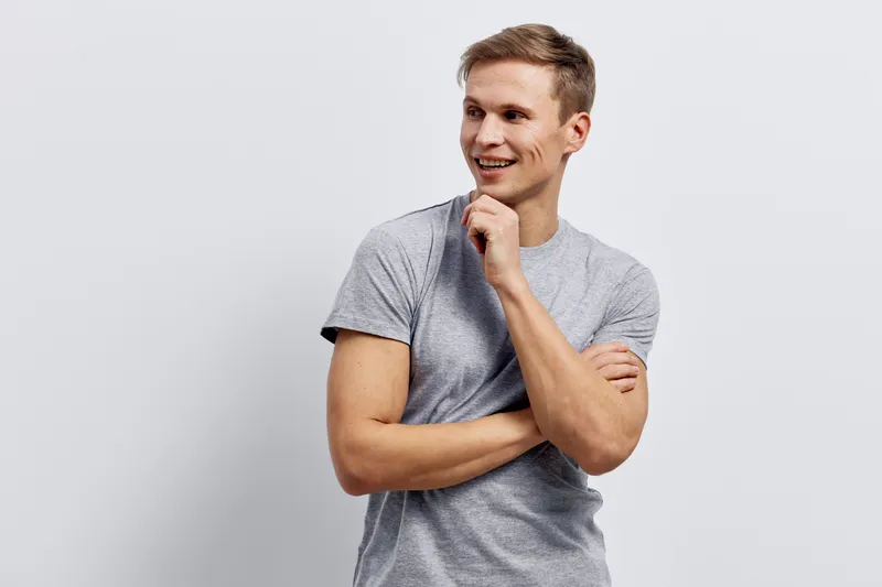 Smiling young man with casual gray t-shirt standing against plain light gray background, expressing happiness, confident pose, cheerful mood, studio portrait, people lifestyle concept, natural facial 