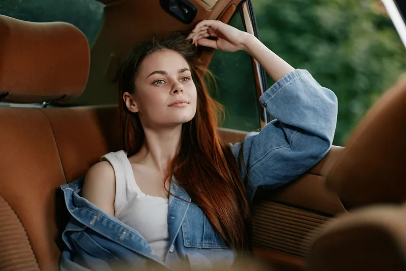 Young woman with long red hair wearing a denim jacket, relaxing in a vintage car, expressing calmness and serenity against a natural green background.