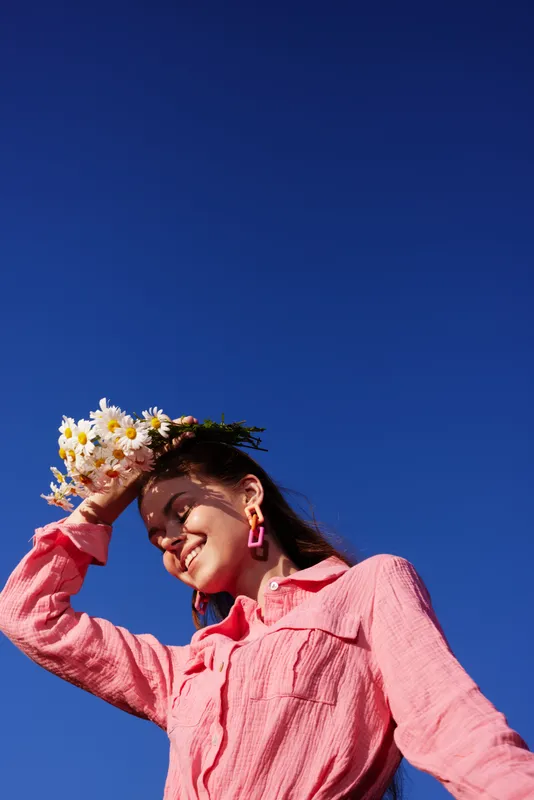 Young woman with a flower crown smiling joyfully in a vibrant pink shirt against a clear blue sky, representing spring and natural beauty