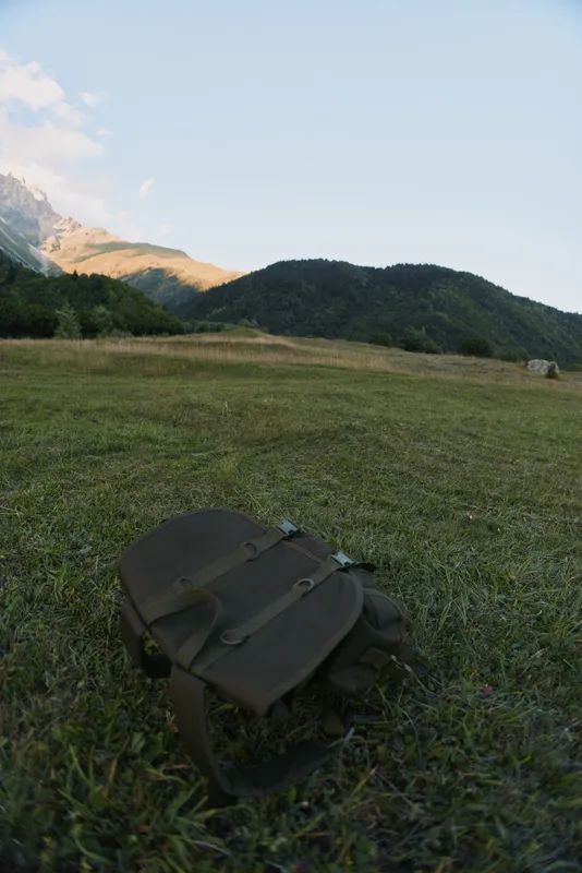 Backpack lying on green grass in mountain valley with forest and hills under clear sky during daytime, outdoor nature scene with travel and adventure concept.