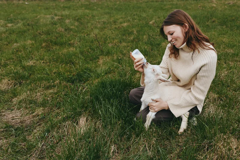 Smiling woman in a cozy sweater feeds a goat kid with a bottle in a grassy field, capturing a warm farm lifestyle moment of care, joy, and connection.