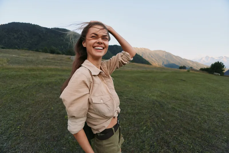 A cheerful woman in a beige shirt stands in a wide field, sunlight on her face, smiling with a hand in her hair. An open landscape conveys carefree outdoor mood.