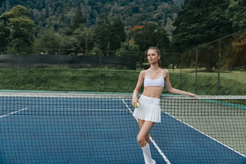 Young female tennis player in white sportswear poses confidently on a court, showcasing athleticism and determination amidst a lush green backdrop
