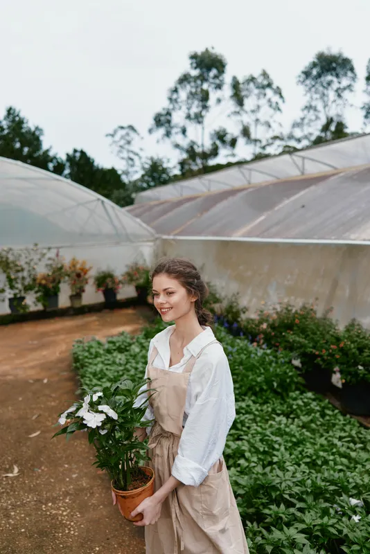 Young woman in garden with potted plant, wearing casual beige overalls and white shirt, smiling among lush greenery in a greenhouse setting