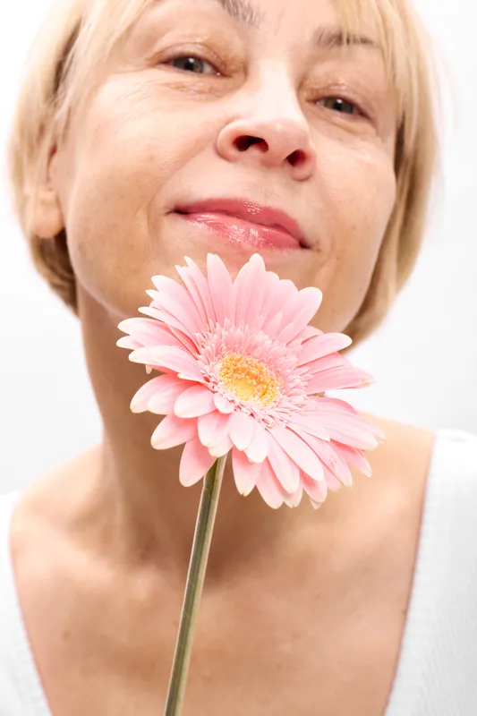 Woman portrait closeup smiling, holding pink gerbera flower near lips, delicate petals in foreground, clean white studio background, spring freshness, beauty and relaxation mood