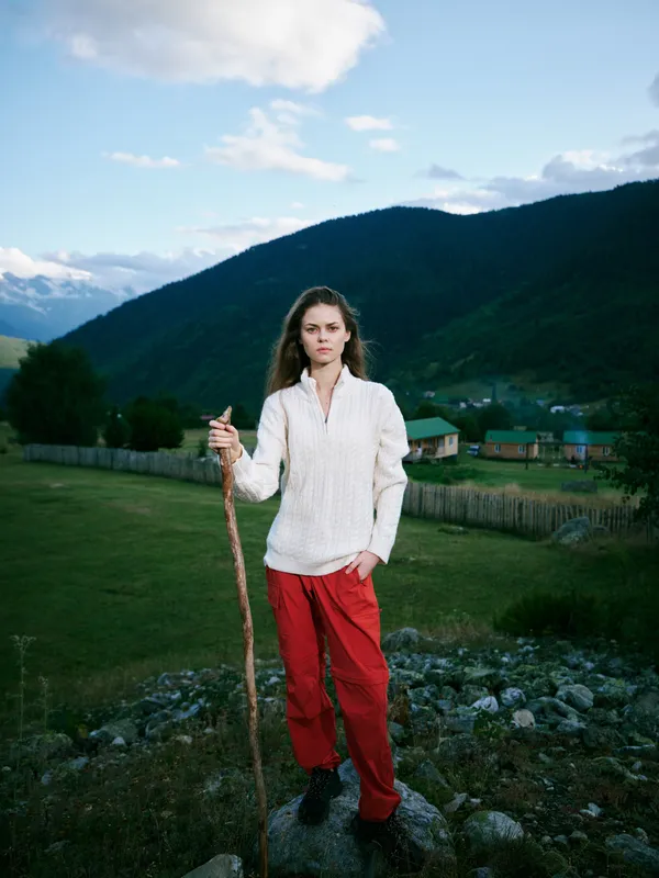 Hiking scene with a woman standing on a rocky overlook, holding a wooden staff, wearing a white knit shirt and red pants, mountains and countryside in the background