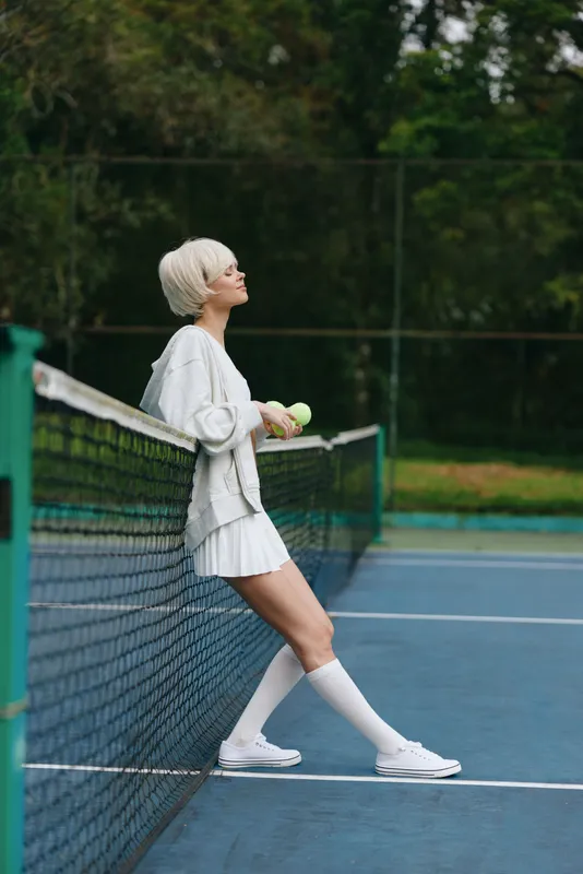 Stylish young woman in a tennis outfit enjoying leisure time on a court, showcasing a relaxed attitude and sporty elegance amidst green surroundings