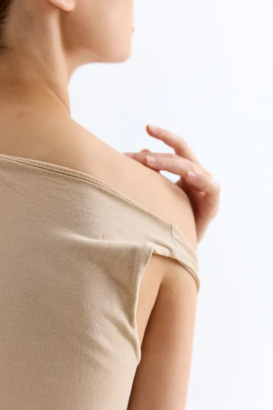 woman touching shoulder with hand, close-up of bare skin, beige top, soft natural light, minimalistic studio background, gentle skincare gesture.