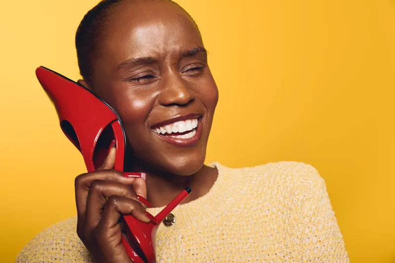 Smiling black woman with dark skin holds red high heel shoe near ear like phone. Yellow background and beige sweater. Concept for creativity, fun, fashion, and joyful emotions in studio.