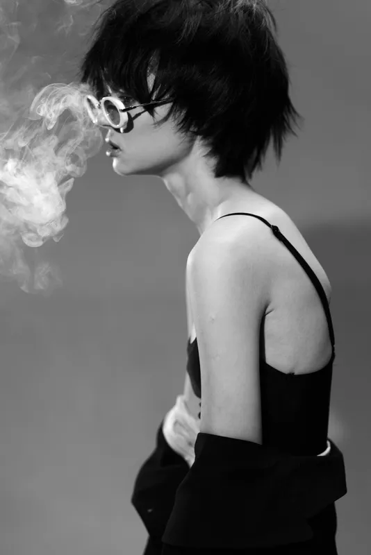 Black and white portrait of a young woman wearing glasses exhaling smoke with a stylish edgy look and short hair.