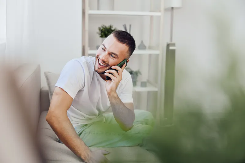 Smiling man engaging in an emotional phone conversation, embodying soft masculinity and mental health awareness, showcasing vulnerability in a cozy home setting.