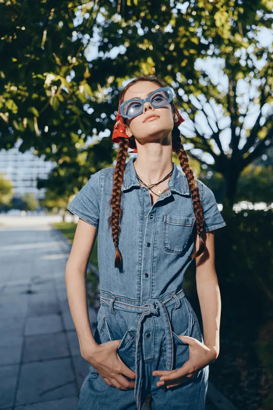 young woman wearing sunglasses outdoors in casual denim dress with braided hair and red bandana under bright sunlight in summer park setting