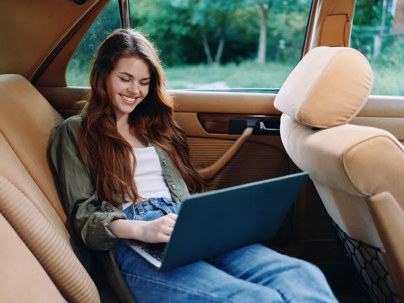 Young woman working on a laptop in a vintage car, showcasing a casual style and joyful emotion, surrounded by a lush green background reflecting a serene atmosphere.