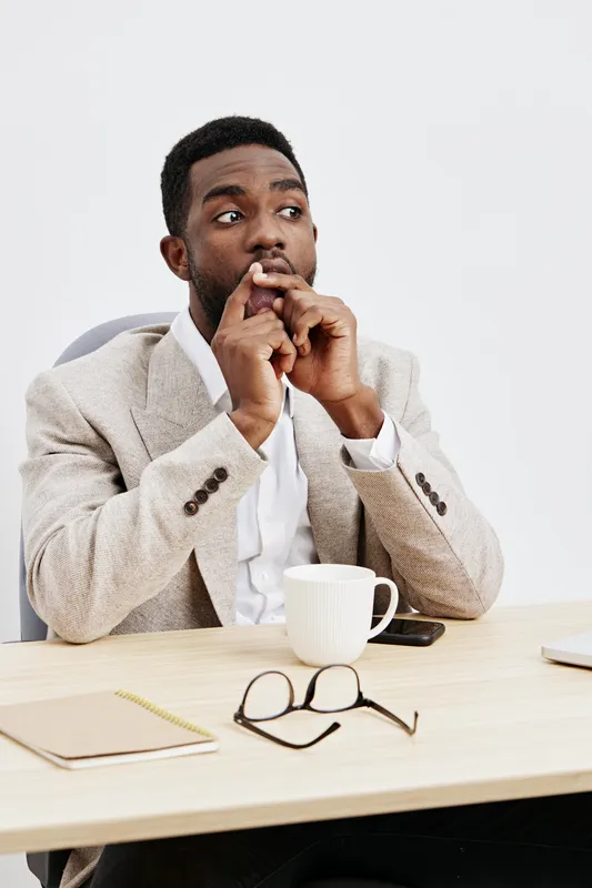 Thoughtful young African American man in formal beige blazer and white shirt, sitting at desk with cup, glasses, notepad, and smartphone, looking away with pensive expression, indoor office background