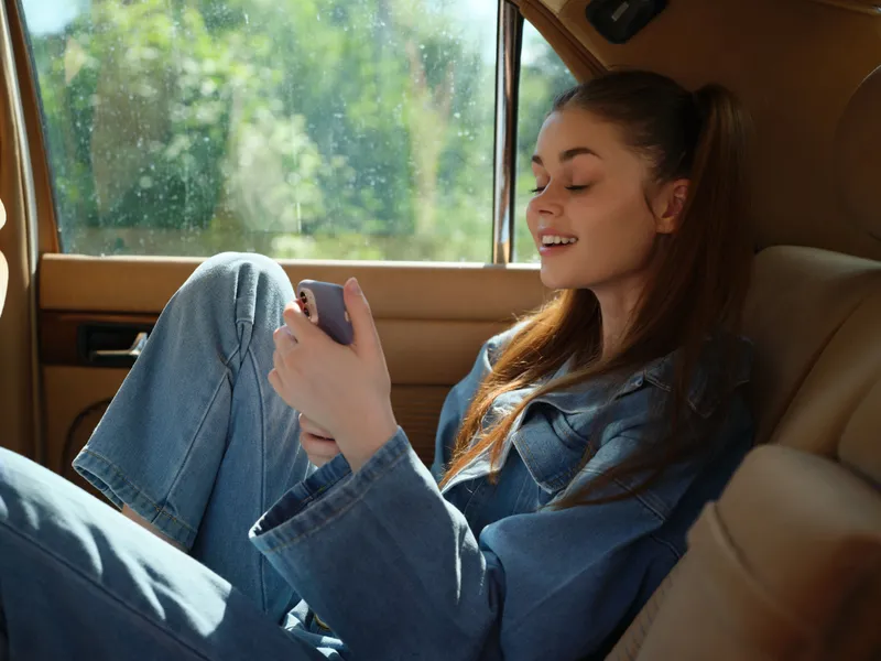 Young woman smiling while using a smartphone in a car, wearing a denim outfit, exuding joy and relaxation in a bright and natural setting.