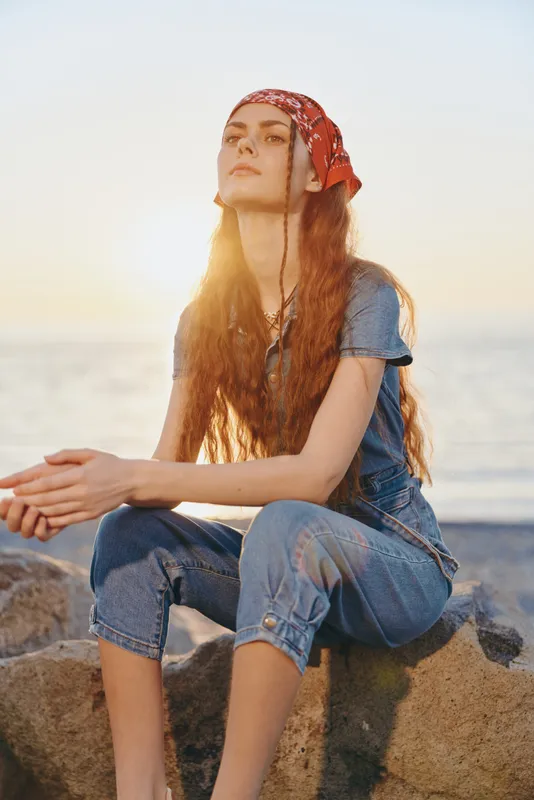 Lifestyle woman in modern boho-western denim street style wearing a red bandana, relaxed pose by the water at sunset, film color warm tones enhancing calm mood.