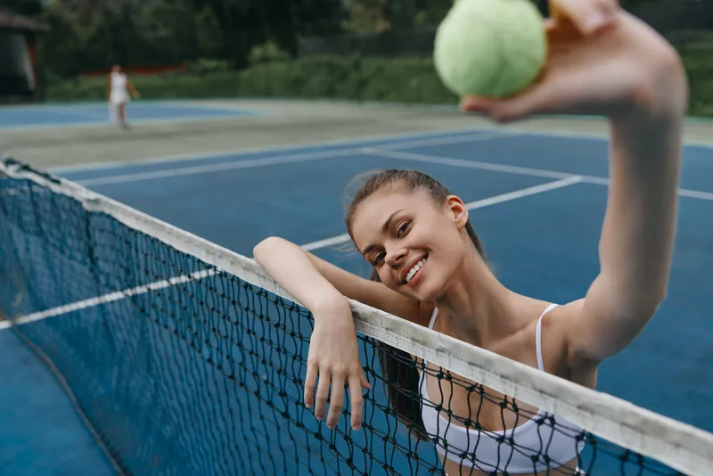 Young female tennis player smiling and holding a tennis ball, showcasing athleticism and joy on a blue outdoor court, perfect for sports and fitness concepts