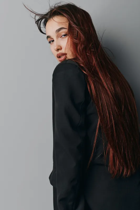 Woman with long brown hair and natural makeup wearing black jacket posing against gray wall. Portrait of young female model with serious expression and casual hairstyle in studio setting.