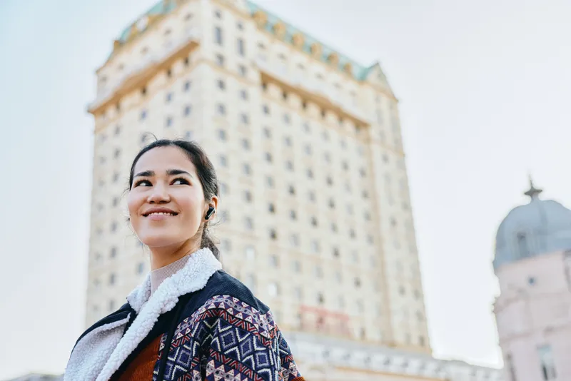 Smiling woman wearing a patterned jacket stands in front of a tall city tower, urban landscape and travel mood, street style, daylight photography, confident expression, candid urban vibe