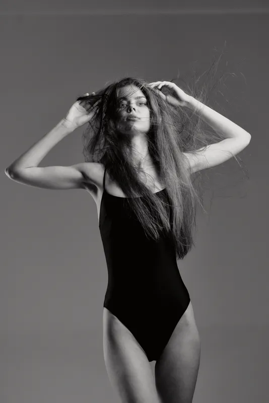 Black and white portrait of a woman with long hair wearing a black swimsuit, posing confidently with hands in her hair.