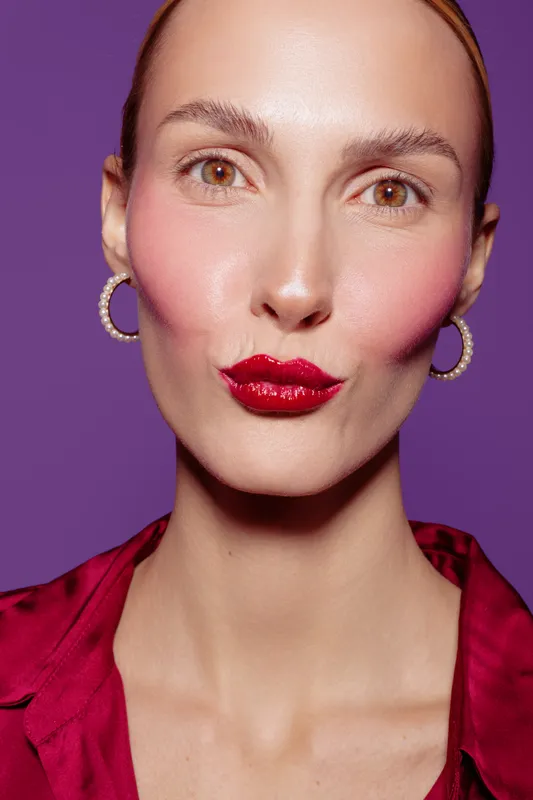 Close-up of a woman with red lipstick and pink blush making a kiss face on purple background. Concept of beauty, makeup, fashion, and confident female portrait.