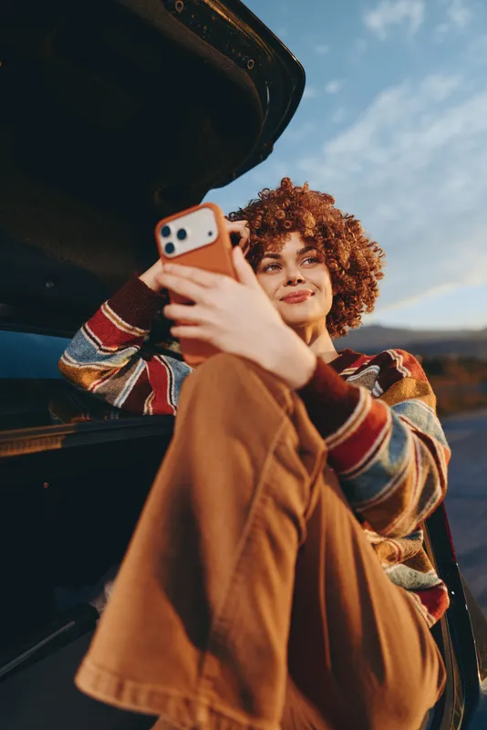 Cheerful woman outdoors taking selfie with smartphone, curly hair and warm smile, leaning by car door at golden hour, casual jacket, natural light, scenic background, lifestyle moment today