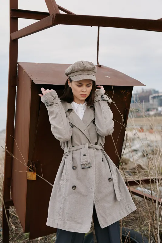 Young woman in textured grey coat and matching cap stands outdoors near rusty metal structure. Autumn fashion concept with moody natural background and casual urban style clothing.