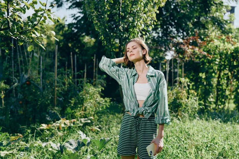 Woman outdoors in a sunny garden stands relaxed with hands in pockets, wearing a striped shorts and white top, enjoying nature, a silent smile, casual fashion, fresh air, carefree mood.