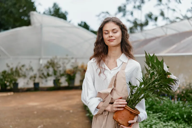 Young woman in a greenhouse holding a potted plant, wearing casual attire, exuding a serene expression, surrounded by greenery and natural light