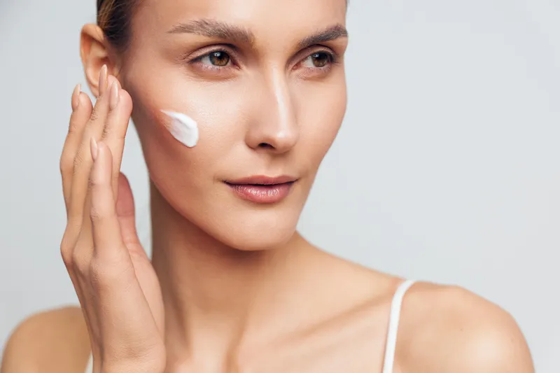 Woman skincare face cream applying closeup beauty hydration moisturize wellness, white cream on cheek, soft studio lighting, neutral background, calm expression, smooth skin